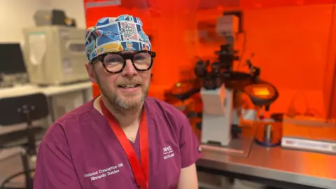 Head and shoulders image of embryologist Phillip Milne who is wearing a burgundy hospital scrubs and a multi-colour surgeon's cap. He's looking at the camera. He has a redish greying beard and moustache and is wearing thick tortoise-shell rimmed glasses.