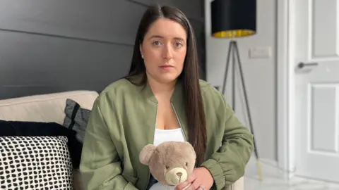 BBC Victoria Buckley is wearing a white top and khaki green bomber jacket. She has long dark brown hair and is looking directly at the camera with a sad expression. She is holding a brown teddy bear that says "Ollie" on its jumper.