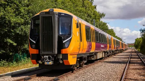 A WMR Class 730 train can be seen on tracks. It has several carriages with orange and maroon livery and black front end.
