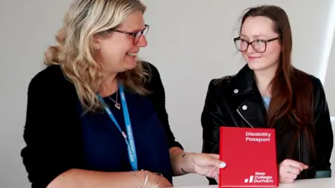 Mhairi Vale is smiling as she holds the disability passport. It is a small book with red covers reading Disability Passport New College Durham. Student Emily Liddell is standing next to Mhairi and smiling slightly at her. They are both wearing glasses. Mhairi has blonde wavy hair and is wearing a navy blouse and black cardigan. Emily has long dark red straight hair and is wearing a black leather jacket and light blue t-shirt. 