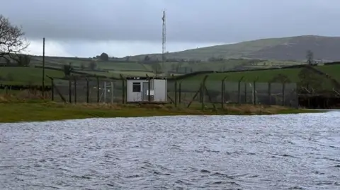 BBC Weather Watcher Gordy Bingham Image of water beside land at Katesbridge in County Down. There is a hut surrounded by a fence and hills in the background.