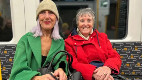 A woman with lilac hair and a beige beanie hat wearing a green coat sitting next to a woman with a red coat and short grey hair.