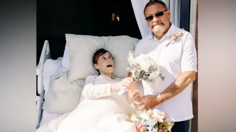 Karen and Stephen Glossop dressed in white holding flowers in a hospice bed
