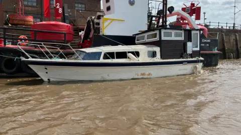 A small white boat in the foreground and a larger black vessel behind it. The smaller boat has a black shed attached to the deck.