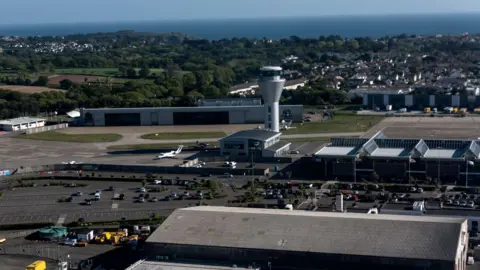 BBC Jersey Airport photographed from a drone on a clear day. The air traffic control tower is visible, as are aircraft hangars and the car park. The sea is visible in the far distance.