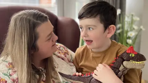 A young boy is sitting on his mother's knee. He is holding a toy dinosaur. He has dark brown hair and is wearing an orange jumper. His mother has blond hair and is wearing a floral blouse.