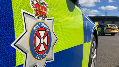 Close up shot of a police car with blue and yellow markings, and a grey Wiltshire Police badge.