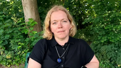 Chloe Purcell is wearing a black shirt with a necklace and is sitting on a bench in front of green shrubs and a tree