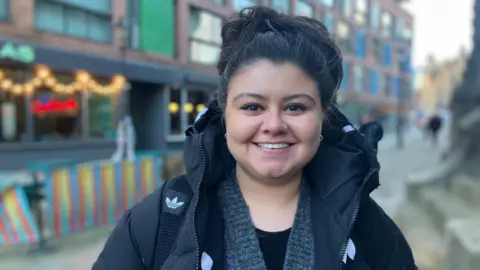 Elizabeth Baines/BBC A woman with black hair tied up on top of her head smiles. She wears a back puffer coat. Behind her, Leeds is blurred.