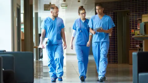 Three junior doctors walking down a hospital corridor while wearing blue scrubs