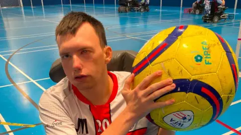 A man wearing a white and red sports top is holding a yellow ball with blue and red stripes. He is looking at the camera whilst sitting in a wheelchair inside a sports hall with a blue floor.