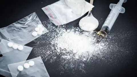 Snap bags containing small white pills, alongside a spoon, and syringe, lying alongside a small pile of white powder.