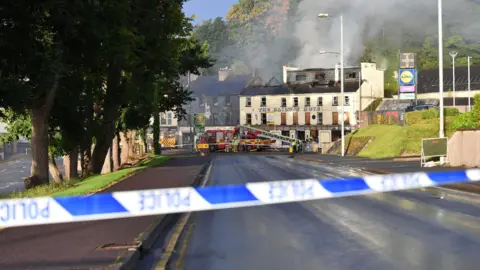 Pacemaker Police tape in the forefront of the image. In the background is the derelict Railway Hotel in Enniskillen. There are firefighters and a fire engine outside. There is smoke coming out of the top of the building and fire damage is visible on the roof. 