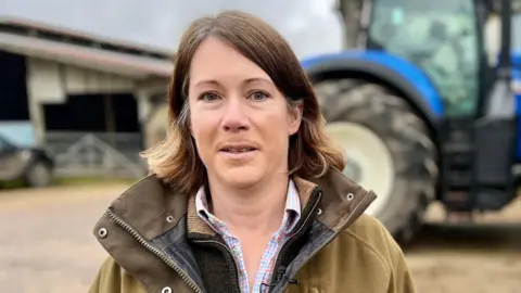 Claire Wright stands in a green winter coat in a farmyard, in front of a large blue tractor.