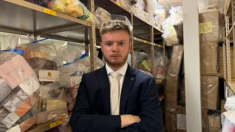 A man in a black suit with a beige tie. He looks fairly cross and is standing in a room full of large transparent plastic bags. The bags are overflowing with seized goods. They are on shelves either side of where he is stood. 