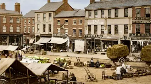 Edwin Williams The restored image of Preston Market Square showing a row of shops with cloth awnings in the background and stalls, carts and wheeled trolleys in the foreground, but coloured in shades of brown and green 