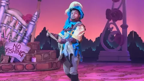 Zak is wearing a blue and purple panto outfit and is stood on stage with a purple painted background and some large props and painted effects. He has a blue hat on with a long feather coming out from it.
