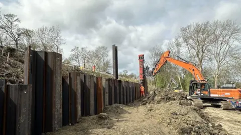 Network Rail A large orange digger working to dig dirt. There are trees in the background.
