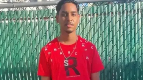 Family A young man in a red t-shirt and a silver chain with the letter s on, looking at the camera