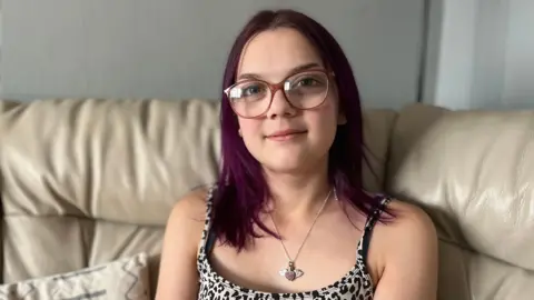 BBC A teenage girl with glasses, purple hair and a heart necklace sitting on a sofa and smiling at the camera