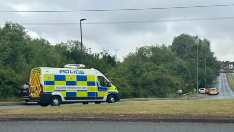 A police van is parked on a grass roundabout. Ahead are three cars including a police car in a crashed position on a road. 