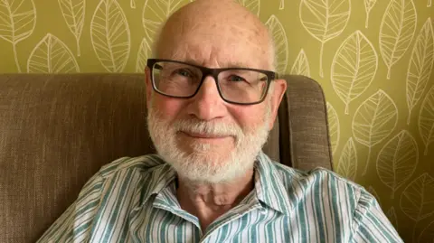 Malcolm Pemble sitting in an armchair is wearing a striped shirt, and dark framed glasses. He has blue eyes and a white beard.