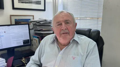 BBC/Kate Bradbrook Man with short grey hair and moustache sits at a desk with a computer monitor in the background. He wears a blue and white striped shirt.
