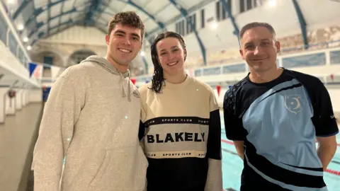 Left to right standing in front of the pool: Tyler, a young man earing a grey hoodie, Amy, a young woman in a blue, white and grey sweater and Dave, a man dressed in a blue t-shirt. All smile at the camera.