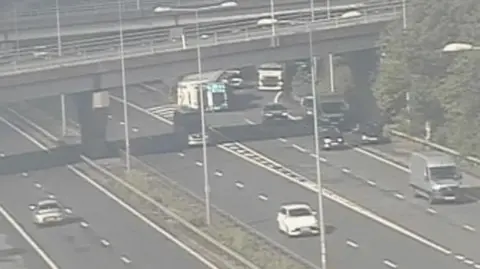 An overturned lorry can be seen on the right hand side near the end of a slip road. Traffic is passing it in another lane. 