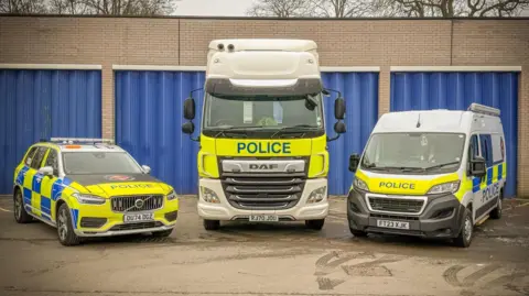 A police lorry with a police car on the left and a van on the right. They are parked in a row, with their bonnets facing the camera, outside a garage with several blue folding doors, which are closed. 