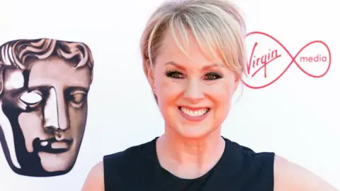 PA Sally Dynevor in a black dress, smiling, with one hand on her hip, poses on the red carpet.
