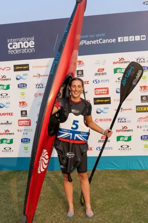 David McBay/Paddle UK A woman in watersports kit, holding a canoe and paddle. She has brown hair in two plaits and is smiling and looking at the camera. Behind her is a board with lots of businesses' logos on it.