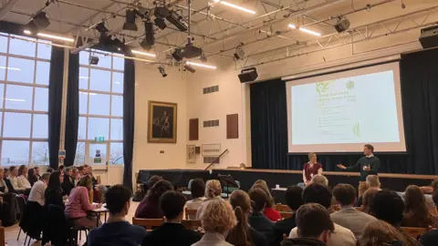 Alex Osborne/ BBC A brightly-lit school hall, with rows of students listening to a presentation by two teachers, the 'Great Guernsey Climate Change Debate 2026' featuring on a projector 
