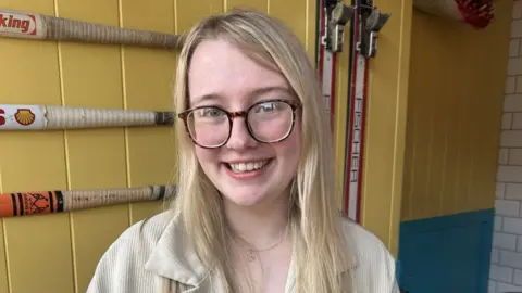 A woman with long blonde hair and brown glasses in sat inside a building with yellow walls. She is smiling at the camera