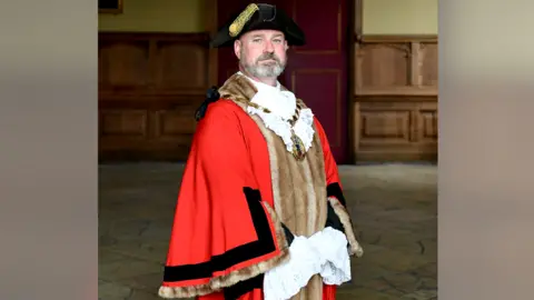 KLWNBC Current mayor of King's Lynn Steve Bearshaw wearing red ceremonial robes. He has a black hat with a gold badge. He wears heavy gold chains around his neck. The robe is lined with brown fur. 