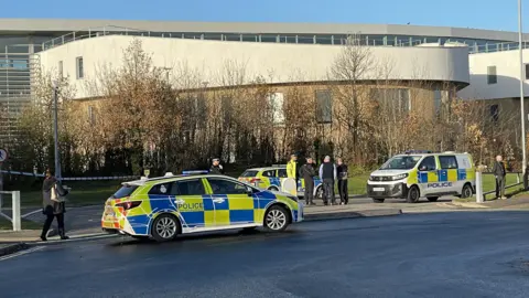 Zoie O'Brien/BBC Two police cars and a van are parked on a road. A police cordon has been set up across part of the road and in front of a large college. Officers speak with people in the area. It is a sunny day.