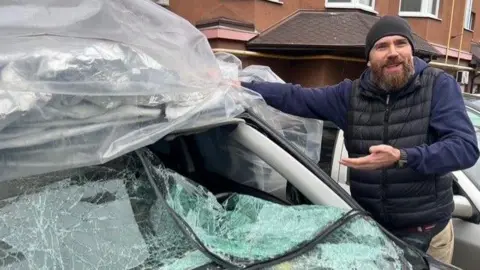Oleksandr Vilko, who has a beard and is wearing a black vest and beanie, points to his car, which has a shattered windshield. 