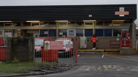 BBC/NEIL OXLEY The exterior of Royal Mail's Washington Delivery Office. There are two red vehicles parked behind red fence railings. There is an unidentifiable postal worker standing next to an empty crate.