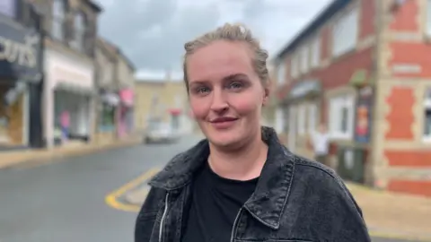 Gemma Dillon/BBC A lady is stood at a t- junction with her hair tied back wearing a black denim jacket with a black t shirt underneath.
Behind her is a row of shops on the left hand side and a red brick public house to her right.