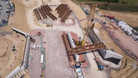 National Highways An aerial shot of the area around the bridge construction, with a large yellow crane and several massive steel beams which dwarf the cars and vans parked around the site. 