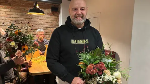 Stuart Leatherbarrow, who is bald and has a short grey beard, stands in front of a table where men are arranging flowers. He is wearing a black hoodie and holding a large beautiful bouquet.