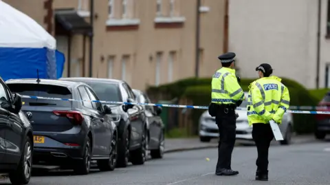 A male and a female police officer standing in front of a line of blue and white police tape blocking off a street. Parked cars, homes and a blue and white forensic tent can be seen on the left of the image.