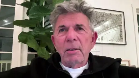 BBC A man in his 50-60s is speaking to camera during a video call. Behind him on the wall is artwork and a plant pops out behind his left shoulder. He's wearing a dark hoody and a white t-shirt