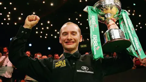 Graeme Dott after winning the World Snooker Championship in 2006. He is holding his trophy in the air. It is silver and has green ribbons attached.