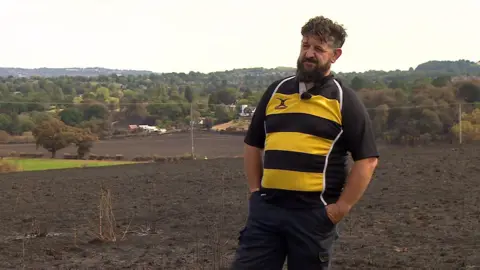 A man wearing a yellow and black top stands in a charred field with his hands in his pockets.