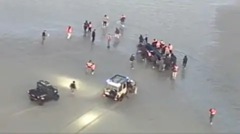Two cars in the shallow water with people surrounding what appears to be a dingy boat with people inside the vessel