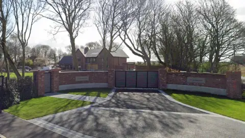 A garden wall with a gate and a sign saying "Otterbrook". In the background is a large, detached property in extensive grounds.