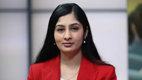 Zarah Sultana has long black hair and a red jacket. She wears white earrings and looks at the camera.