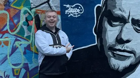 Mark Dallas/Instagram Mark Dallas next to a mural painted of himself dressed in a white The North Face quarter zip.