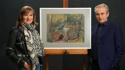 BBC Studios A man and a woman standing in front of a framed painting on an easel. She is wearing a black leather jacket and a patterned scarf. He is wearing a dark blue shirt and a blue suit jacket.